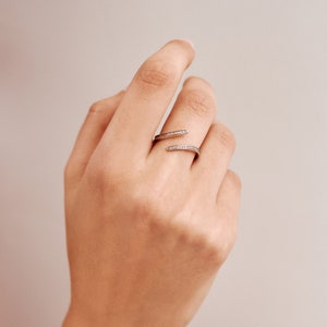 Pu&ograve; includere: Un anello avvolgente color argento con un design diviso. L'anello presenta una fila di piccole pietre preziose trasparenti lungo le fasce superiore e inferiore. L'anello &egrave; indossato su una mano con uno sfondo neutro.