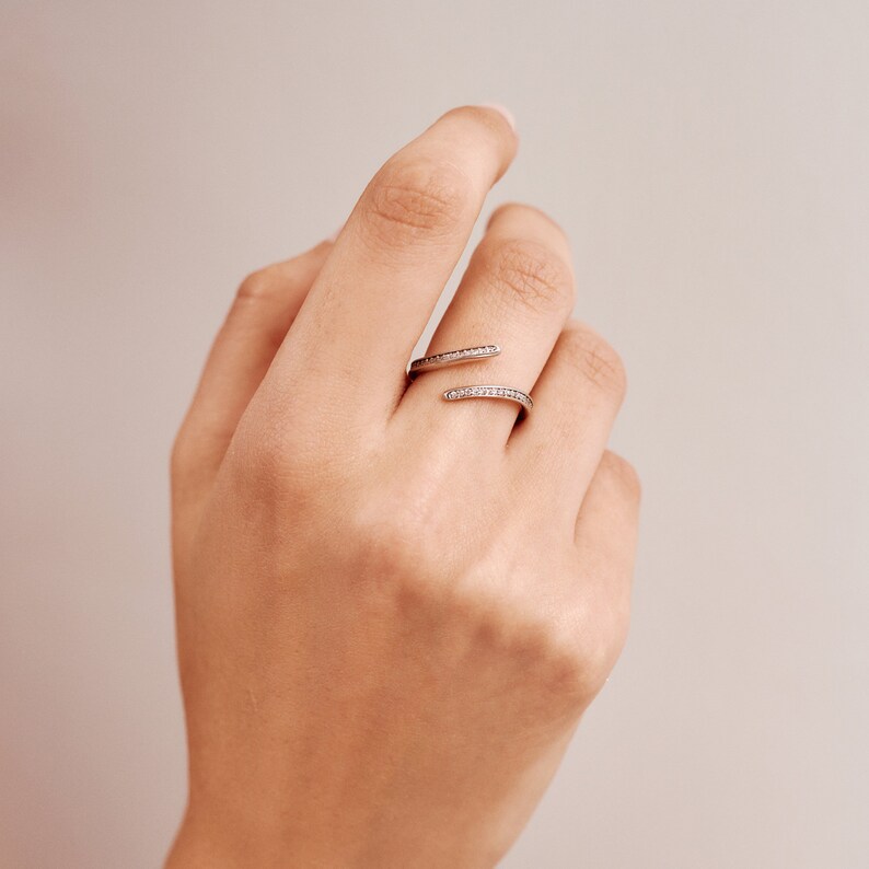 Pu&ograve; includere: Un anello avvolgente color argento con un design diviso. L'anello presenta una fila di piccole pietre preziose trasparenti lungo le fasce superiore e inferiore. L'anello &egrave; indossato su una mano con uno sfondo neutro.