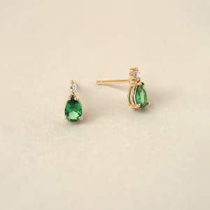 May include: A pair of gold stud earrings featuring a pear-shaped green gemstone suspended below a small, round, clear white gemstone. The earrings have a post-back closure and are crafted with a polished gold metal setting for a refined appearance.