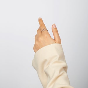 Pu&ograve; includere: Un anello avvolgente color argento con piccole pietre trasparenti. L'anello &egrave; su una mano con una manica color neutro. L'anello si avvolge attorno al dito, creando un design moderno e minimalista. Lo sfondo &egrave; bianco.