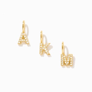 May include: Three gold letter charms shaped as A, K, and M, each featuring a small attached jump ring at the top. The surface of each letter is encrusted with small, clear, sparkling stones that catch the light, creating a bright, reflective finish.