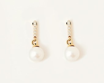 May include: A pair of gold drop earrings featuring a vertical bar set with small clear stones, connected to a round white pearl. The earrings have a polished gold finish and a simple, elegant design that hangs vertically from the top attachment point.