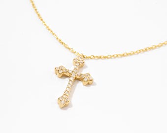 May include: A gold chain necklace featuring a cross pendant adorned with small, clear, sparkling stones. The cross has a decorative, flared design at each of its four ends, and the entire piece is crafted from a polished, reflective gold-tone metal.