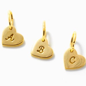 May include: Three gold-colored heart-shaped charms, each featuring a single engraved letter. The left charm displays the letter A, the center charm displays the letter B, and the right charm displays the letter C. Each charm includes a small jump ring.