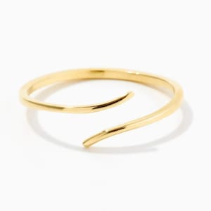 May include: A polished gold-tone metal ring featuring an open-ended bypass design. The band has a smooth, rounded surface that tapers slightly at the two ends, which overlap without touching, creating a continuous circular silhouette.