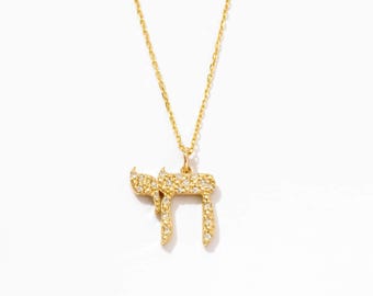 May include: A gold-toned necklace featuring a pendant shaped like the Hebrew letter Chai, which is encrusted with small, clear, sparkling stones. The pendant hangs from a delicate, matching gold-toned chain with a simple, refined link design.