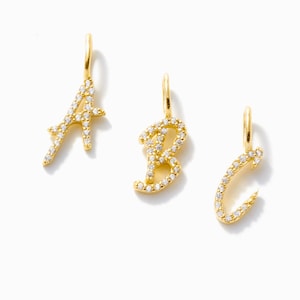 Könnte beinhalten: Drei goldfarbene Initialen-Anhänger, die Buchstaben A, B und C, jeweils mit kleinen, klaren Edelsteinen besetzt. Die Anhänger haben oben eine Schlaufe zum Aufhängen und werden vor weißem Hintergrund präsentiert.