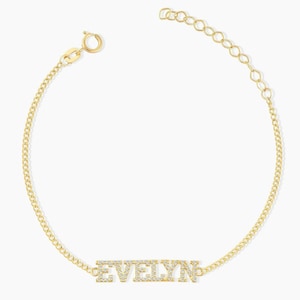 May include: A gold-toned chain bracelet featuring a central nameplate with the name EVELYN spelled out in clear, sparkling stones. The bracelet includes a delicate link chain, a circular spring ring clasp, and an adjustable extender chain.