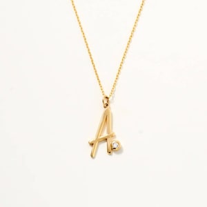 May include: A gold-toned necklace featuring a delicate chain and a pendant shaped like the capital letter A. The letter pendant is accented with a single, small, round, clear gemstone set into the lower right side of the character's structure.