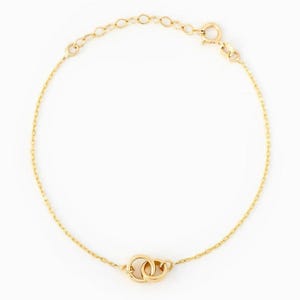 May include: A delicate gold chain bracelet featuring two small, interlocking circular rings as a central focal point. The bracelet includes a thin, polished gold link chain and a secure circular spring ring clasp with an adjustable extension chain.