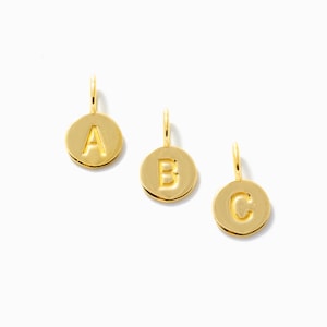 Puede incluir: Tres colgantes redondos dorados, cada uno con una letra diferente: A, B y C. Las letras están grabadas en los colgantes. Cada colgante tiene un pequeño lazo en la parte superior para colgar. Los colgantes están sobre un fondo blanco.