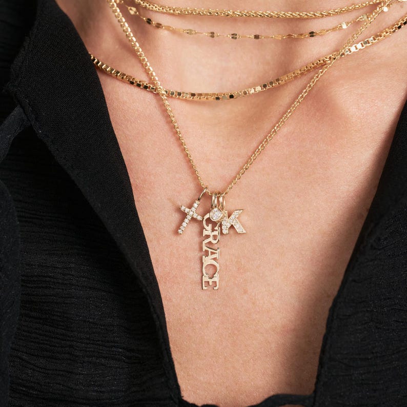 Pode incluir: Um colar de ouro com v&aacute;rios pingentes, incluindo uma cruz, uma letra K e um pingente vertical com a palavra "GRACE" escrita em diamantes.