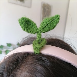 Può includere: Un cerchietto verde all'uncinetto a forma di pianta con due foglie. Il cerchietto è indossato sulla testa di una persona.