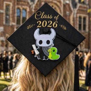 May include: A black graduation cap with gold text reading "Class of 2026" and a cartoon character design. The cap is worn by a person with long, wavy blonde hair, and a black graduation gown. The background features a brick building and other graduates.
