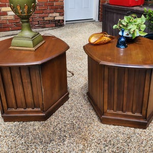 May include: Two hexagonal wooden end tables with a medium brown finish. Each table features vertical slat detailing on the sides and a flat top surface. One table supports a green lamp, while the other holds a small amber glass dish and a blue bell.