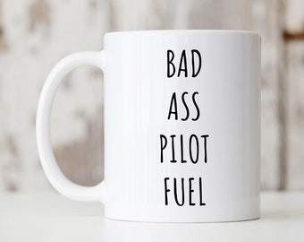 以下が含まれることがあります： 黒い文字で「Bad Ass Pilot Fuel」と書かれた白いセラミック製のコーヒーカップです。