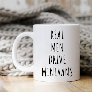 May include: White ceramic mug with the text "REAL MEN DRIVE MINIVANS" in black, displayed against a neutral background. The mug has a curved handle and is likely used for beverages. The overall theme is novelty and humor.
