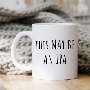 Può includere: Tazza in ceramica bianca con testo nero che recita "This may be an IPA". La tazza è appoggiata su una superficie di legno con una coperta di lana bianca sullo sfondo.