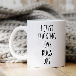 May include: White ceramic coffee mug with black text that reads "I just fucking love bugs ok?" The mug is sitting on a wooden surface with a white knitted blanket in the background.