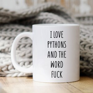 May include: White ceramic mug with the text "I LOVE PYTHON'S AND THE WORD FUCK" in black. The mug is set against a neutral background, with a soft, knitted blanket.