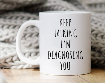 May include: White ceramic coffee mug with black text that reads "Keep Talking I'm Diagnosing You". The mug is sitting on a wooden surface with a white knitted blanket in the background.