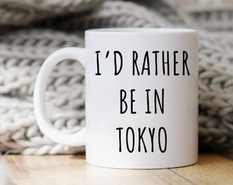 May include: White ceramic mug with black text that reads "I'd rather be in Tokyo". The mug is sitting on a wooden surface with a white knitted blanket in the background.