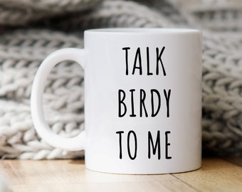 Op de afbeelding: Witte keramische mok met zwarte tekst die "Talk Birdy To Me" zegt. De mok staat op een houten oppervlak met een wit gebreid deken op de achtergrond.