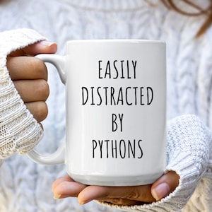 May include: White ceramic coffee mug with black text that reads "Easily Distracted by Pythons".