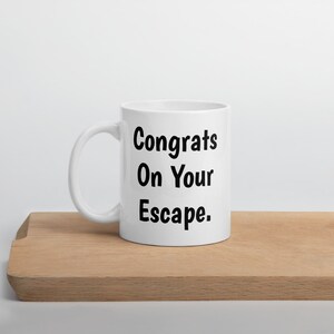 May include: White ceramic coffee mug with black text that reads "Congrats On Your Escape."