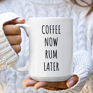 Op de afbeelding: Witte keramische koffiemok met zwarte tekst die "Coffee Now Rum Later" luidt.