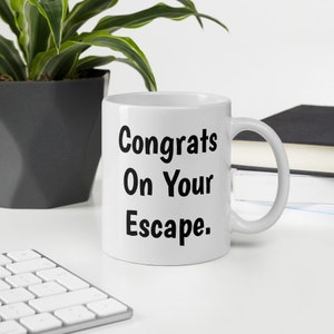 May include: White ceramic coffee mug with black text that reads "Congrats On Your Escape."