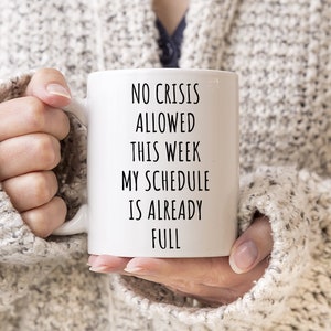 Op de afbeelding: Witte keramische mok met de tekst "NO CRISIS ALLOWED THIS WEEK MY SCHEDULE IS ALREADY FULL" in het zwart. De mok wordt vastgehouden door een persoon die een grijze gebreide trui draagt.