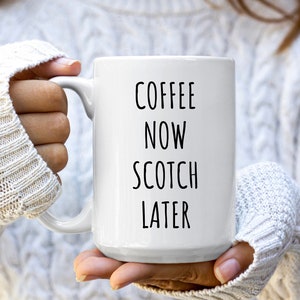 May include: White ceramic mug with the text "COFFEE NOW SCOTCH LATER" in black. The mug is held by a person wearing a white cable-knit sweater. The mug has a large handle.