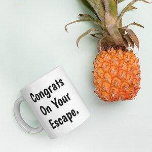 May include: White ceramic mug with black text that reads "Congrats On Your Escape."  A pineapple sits on a light green background.