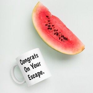May include: White ceramic mug with black text that reads "Congrats On Your Escape." A slice of watermelon is in the background.