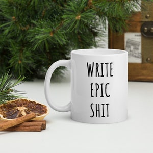 Può includere: Tazza in ceramica bianca con le parole "WRITE EPIC SHIT" in lettere nere e in grassetto. La tazza ha un manico curvo. Lo sfondo comprende vegetazione e oggetti decorativi.