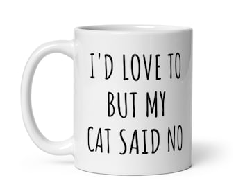 May include: White ceramic mug with the text "I'D LOVE TO BUT MY CAT SAID NO" in black, a humorous statement for cat lovers. The mug has a curved handle and a glossy finish.