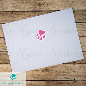 K&ouml;nnte beinhalten: Ein wei&szlig;er Umschlag mit einem rosa Pfotenabdruck-Aufkleber mit einem Herz in der Mitte. Der Text "The Elysian Creations" befindet sich am unteren Rand des Umschlags.