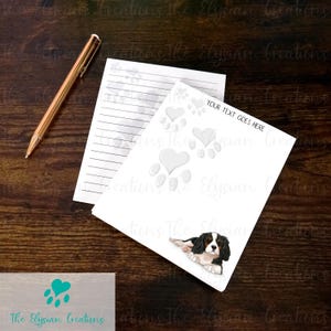 May include: A white notepad with lined paper and a white background with paw prints and hearts. The notepad has the text "YOUR TEXT GOES HERE" printed on it. A black and white Cavalier King Charles Spaniel dog is lying down on the notepad.
