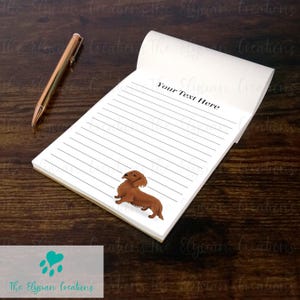 May include: A notepad with lined paper and a brown dachshund illustration. The notepad has the text "Your Text Here" at the top. A gold pen rests beside the notepad on a dark wood surface.