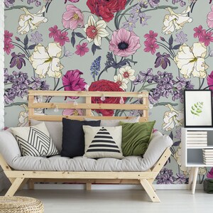 May include: A light gray couch with a floral patterned wallpaper behind it. The couch has a wooden frame and is covered in a light gray fabric. There are four pillows on the couch, two with black and white geometric patterns, one with a black and white striped pattern, and one with a solid green color. There is a small wooden table with a white top and a framed picture on it. The table is next to the couch and has a few books stacked on it. There is a small green suitcase on the floor next to the table.