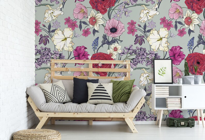 May include: A light gray couch with a floral patterned wallpaper behind it. The couch has a wooden frame and is covered in a light gray fabric. There are four pillows on the couch, two with black and white geometric patterns, one with a black and white striped pattern, and one with a solid green color. There is a small wooden table with a white top and a framed picture on it. The table is next to the couch and has a few books stacked on it. There is a small green suitcase on the floor next to the table.