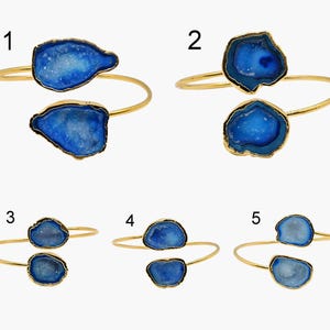 May include: Gold-coloured metal cuff bracelets with two blue geode stone accents. The geode stones have a crystalline interior and are set in a gold-coloured metal frame. The bracelets are displayed against a white background.
