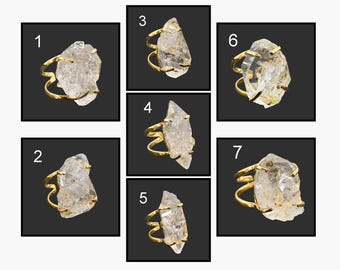 May include: A collection of seven gold-toned rings, each featuring a unique, irregularly shaped, clear crystal stone. The rings are displayed against a dark background, showcasing the natural beauty of the gemstones and the intricate ring designs.