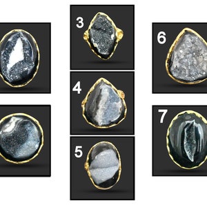 May include: Seven gold-coloured rings, each with a unique, irregularly shaped, dark grey and white geode stone. The stones are set in a gold-coloured metal, with varying oval, round, and teardrop shapes. The rings are displayed against a dark background.