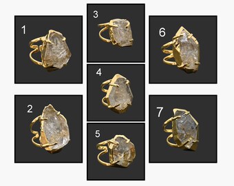 May include: A collection of seven gold-toned rings, each featuring a unique, irregularly shaped, clear crystal stone. The rings are displayed against a dark background, showcasing the intricate details of the jewelry.