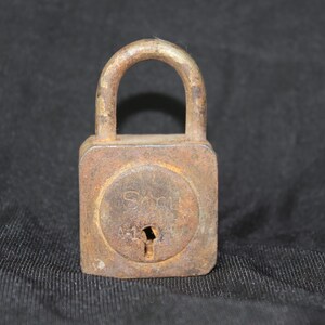 Puede incluir: Un candado oxidado con un cuerpo cuadrado y una cerradura redonda. El candado tiene la palabra "S&M" impresa en él.