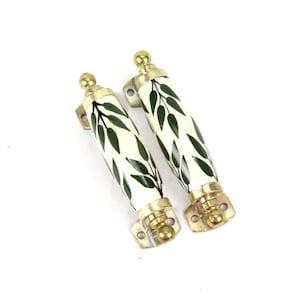 Op de afbeelding: Twee decoratieve kastgrepen. Elke greep heeft een crèmekleurig cilindrisch lichaam met een groen bladerpatroon. De uiteinden en het bevestigingsmateriaal zijn goudkleurig. De grepen zijn ontworpen voor meubels.