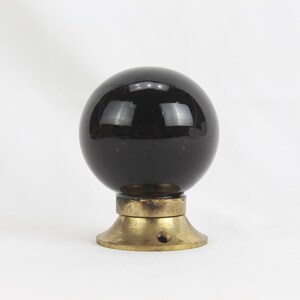 Pode incluir: Um botão de vidro preto com base de latão. O botão é redondo e tem um acabamento brilhante.