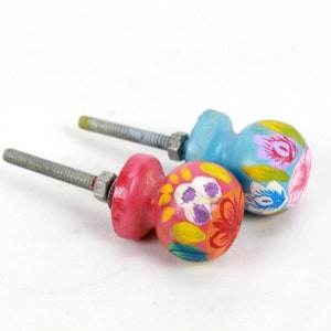 Könnte beinhalten: Zwei bemalte Holzknöpfe mit Blumendesigns. Ein Knopf ist pink mit gelben, orangefarbenen und violetten Blumen. Der andere Knopf ist blau mit gelben, rosa und weißen Blumen.